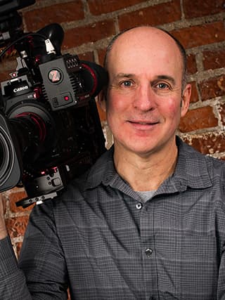 Portrait of David Fitzgibbon smiling with a video camera in front of a brick wall.